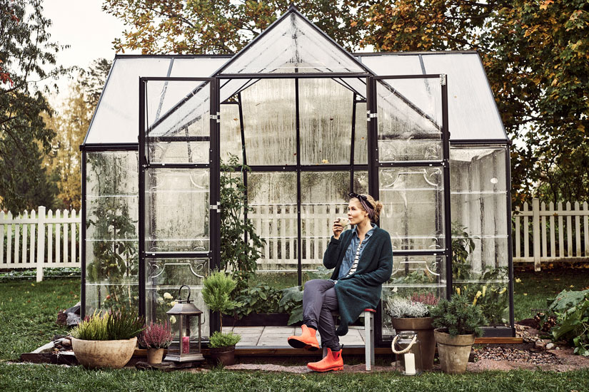 Nainen istuu kasvihuoneen edessä syksyisessä maisemassa juomassa teetä tai kahvia.