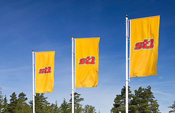 Kolme St1:n lippua liehumassa sinisen taivaan edessä, taustalla puun latvoja.