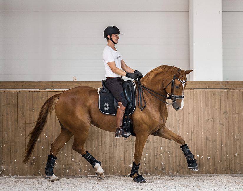 Ville Vaurio ratsastaa ravia valkoisessa T-paidassa hymyillen ruskealla hevosella maneesissa. Hevosella on kankisuitset ja musta satulahuopa